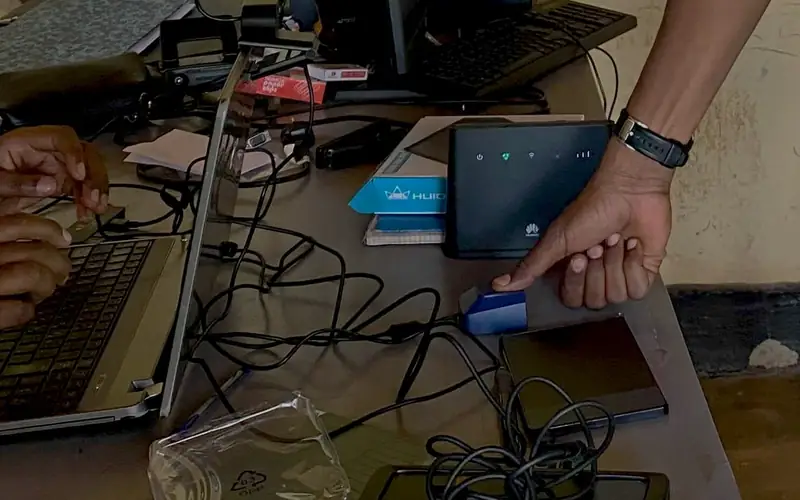 A land records officer presses his thumb onto a fingerprint scanner on a government office desk, beside a laptop where a colleague enters the record