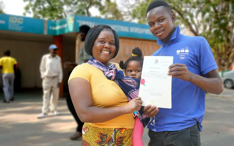 A woman holding her child stands beside a Medici Land Governance field officer, the two of them holding up her newly issued certificate of title